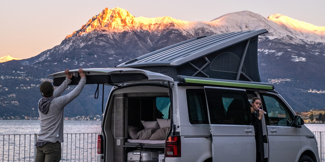 Two people are getting the camper ready for the road by a mountain lake