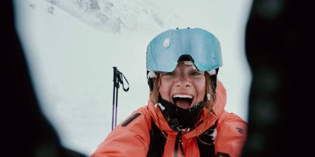 Eine Frau lacht in die Kamera, sie trägt einen Skianzug und hinter ihr ist Schnee zu sehen.