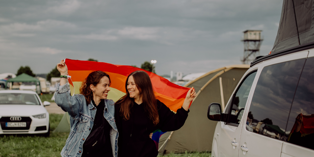 A lesbian couple at a campsite is holding a rainbow flag and smiling at each other.