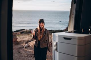 Eine Frau lächelt am Strand nahe dem Camper und hält eine Tasse Tee